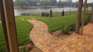 A brick pathway leading to a body of water, surrounded by a fence and greenery.