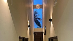 A hallway with a wooden floor, light-colored walls, and a view of palm trees through a window at the end. - GAIA ~ One Bedroom Condo w/Pool. New Build✨ (Puerto Morelos)