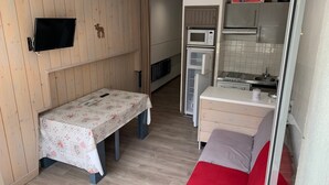 Ein gemütlicher Raum mit einer Holzverkleidung, einem kleinen Tisch mit einer blumigen Tischdecke, einer Bank, einer Küchenzeile inklusive Mikrowelle und Kühlschrank sowie einem an der Wand montierten Fernseher.