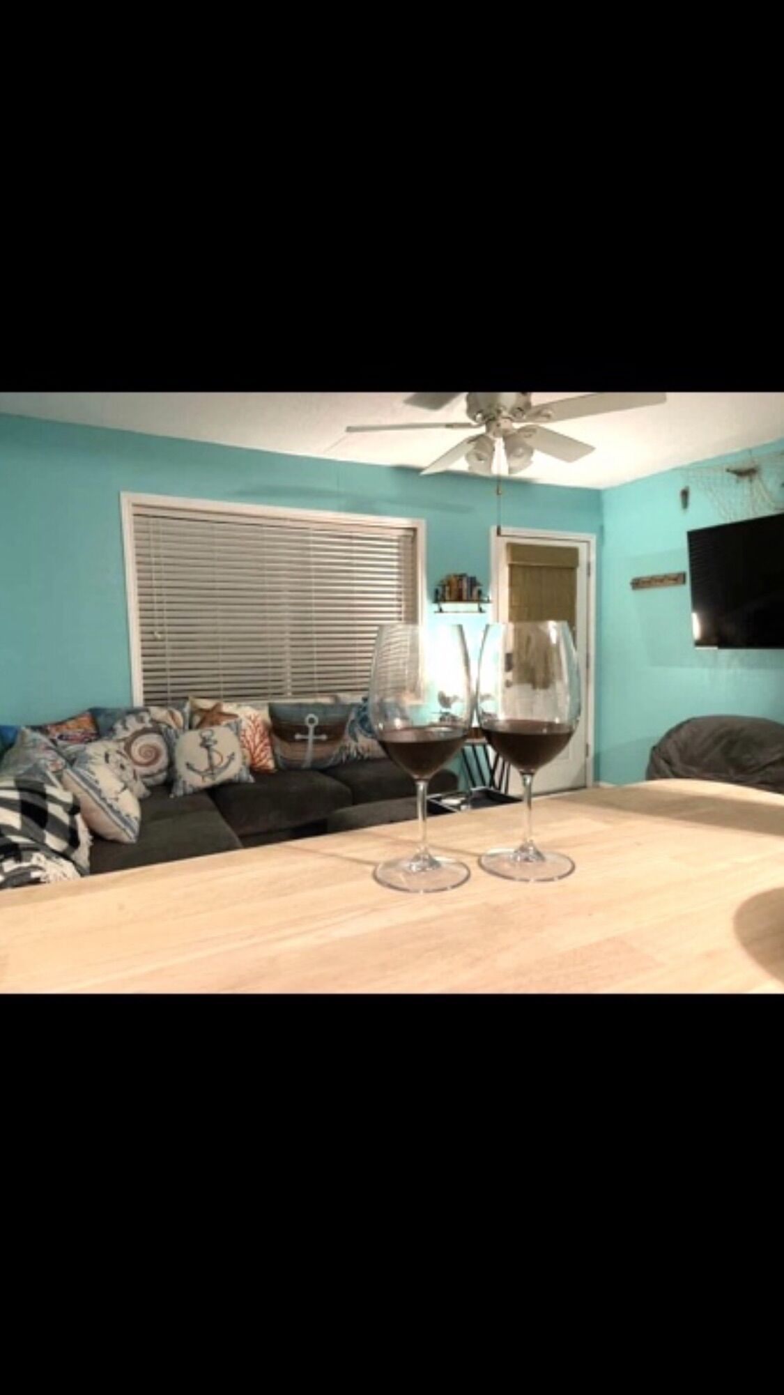A room with teal walls, a ceiling fan, a window with blinds, a sofa with patterned cushions, and two wine glasses on a table.