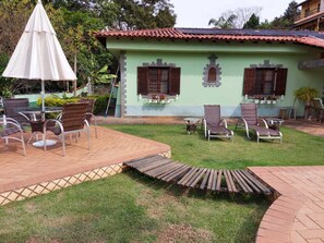 A green building with a tiled roof, outdoor seating area with wicker chairs and a table, and a small garden. - Beautiful farm with stunning views located in a charming and beautiful condominium. (São Roque)