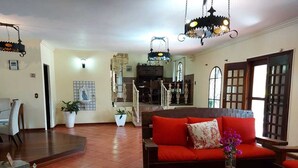 Un amplio salón con un sofá rojo, una mesa de centro y una araña. Al fondo, un comedor con una mesa y sillas.
