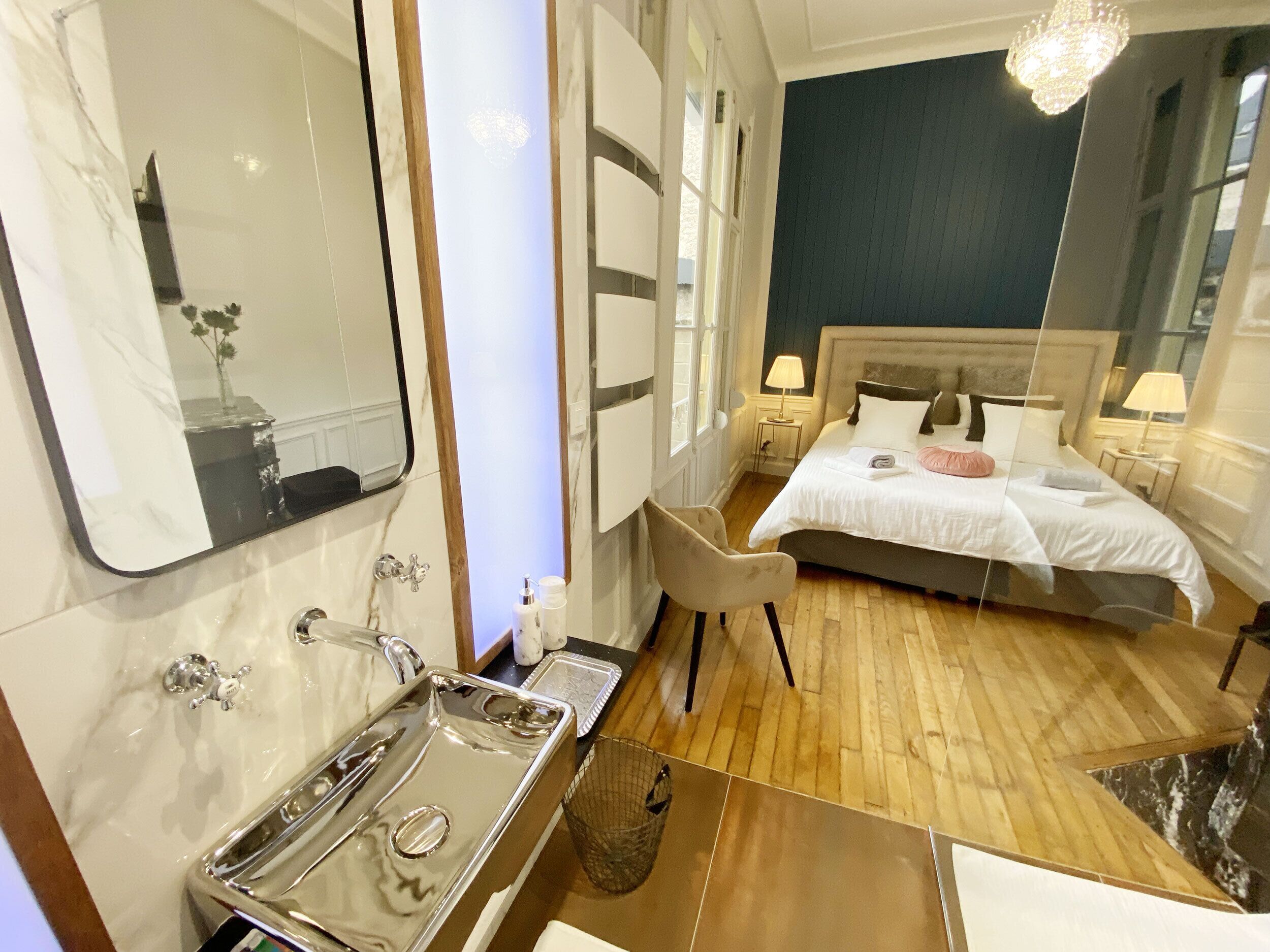 A hotel room with a large mirror, a bed with white bedding, a wooden floor, and a chandelier.