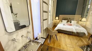 A hotel room with a large mirror, a bed with white bedding, a wooden floor, and a chandelier.