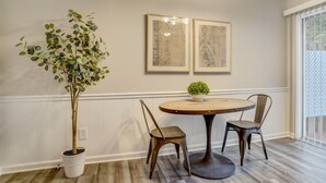 A dining area with a round table, two chairs, a potted plant, and two framed artworks on the wall.