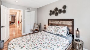 A bedroom with a floral bedspread, a wooden headboard, a wall-mounted decorative piece, and a bedside table with a lamp. - Stunning & Glamorous 3BR-3BA - Sleep 10! (Philadelphia)