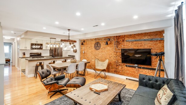 A modern living room with a dining area, a kitchen, and a brick wall with a mounted TV. - Stunning & Glamorous 3BR-3BA - Sleep 10! (Philadelphia)