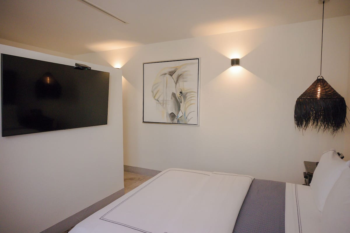 A modern bedroom with a flat-screen TV mounted on the wall, a framed artwork above the bed, and a hanging pendant light.