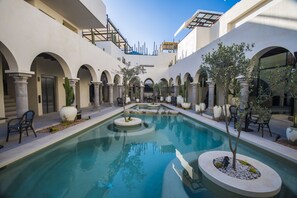 Una piscina rodeada de columnas y arcos blancos, con zonas de estar y plantas en macetas.
