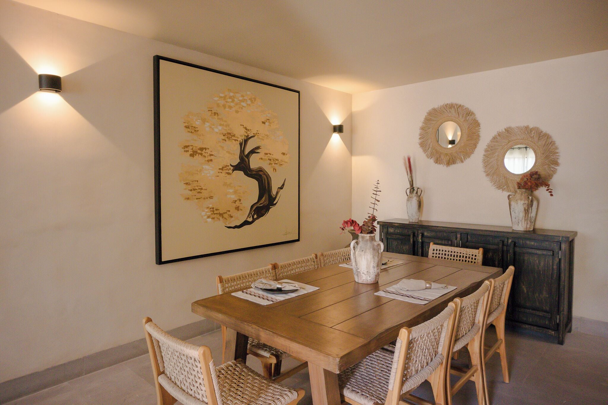 A dining area with a wooden table, wicker chairs, a vase with flowers, and wall-mounted decor.