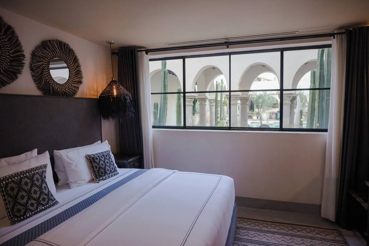 A hotel room with a large bed, a view of an outdoor courtyard with arches, and decorative wall mirrors.