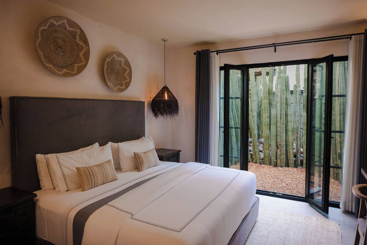 A bedroom with a large bed, a dark headboard, two wall-mounted woven art pieces, a hanging light fixture, and a sliding glass door leading to a cactus garden.