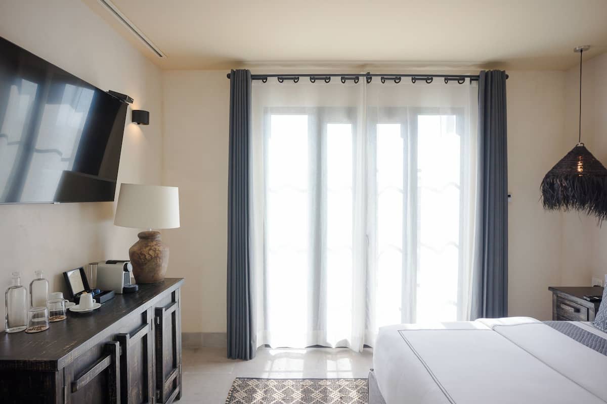 A modern hotel room with a bed, a desk, a TV mounted on the wall, and a large window with curtains.