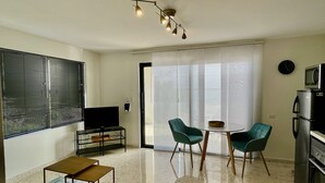 Een moderne woonkamer met een flatscreen televisie, een salontafel, twee stoelen, een bank, een eettafel met stoelen, een koelkast en een klok aan de muur.