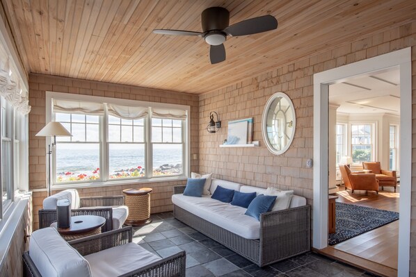 A sunlit room with a wicker sofa, wicker chairs, a ceiling fan, and a view of the ocean through large windows.