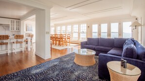 A spacious living area with a large window overlooking the sea, a blue sofa, a glass coffee table, and a dining area with wooden chairs and a white table. - Spectacular 4-Bedroom Luxury Home with Ocean and Mother's Beach Views! (Kennebunk)