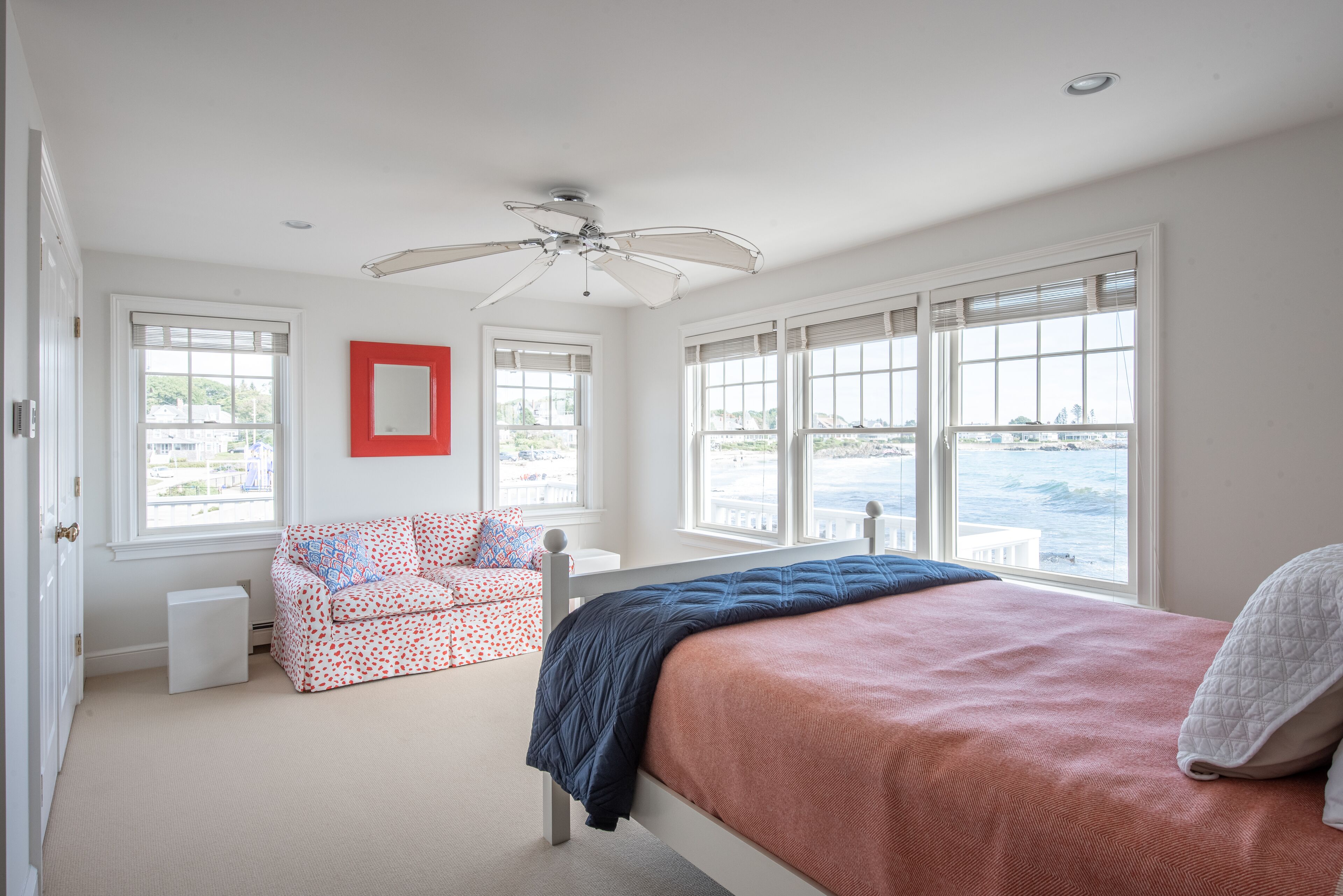 A bedroom with a bed, a window with a view of the sea, a ceiling fan, and a red-framed mirror.