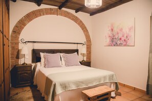 Un dormitorio con un arco de ladrillo, un cabecero de madera, una cama con ropa de cama blanca, dos mesitas de noche con lámparas y un banco de madera.