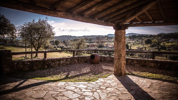 Un patio empedrado de piedra con una pérgola rústica de madera, que da a un paisaje rural con árboles y colinas.