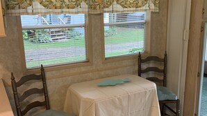 A room with a table and two chairs, a window with blinds, a floral curtain, and a framed sign on the wall. - Peaceful Get-Away Near Conneaut Lake (Harmonsburg)