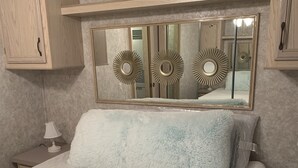 A bedroom with a large bed, a nightstand, a lamp, a mirror with decorative frames, and wooden cabinets. - Peaceful Get-Away Near Conneaut Lake (Harmonsburg)