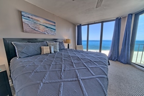 Quarto com uma cama grande, uma pintura de uma praia, mesa de cabeceira com abajur e vista para o oceano através de amplas janelas.