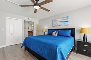 A bedroom with a bed, a ceiling fan, a nightstand, a lamp, a painting, and a door.