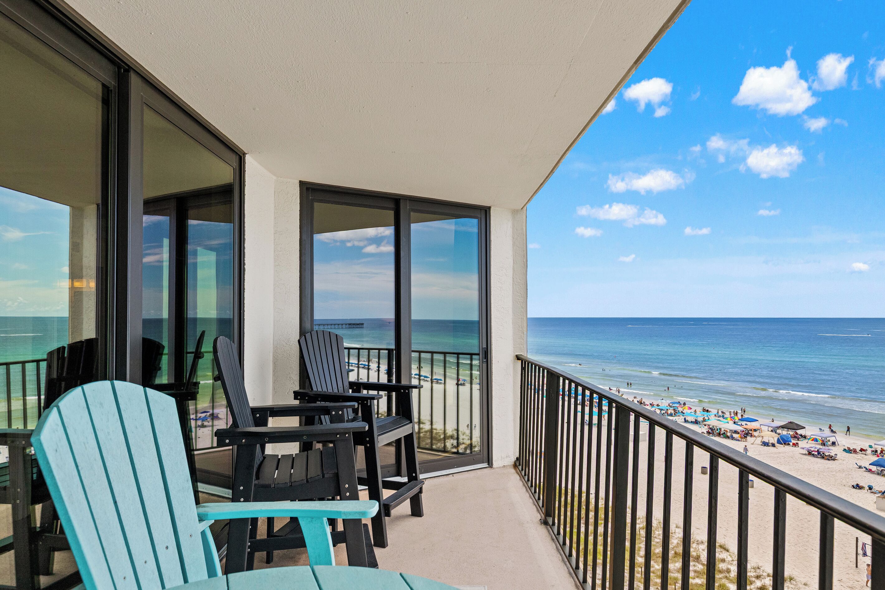 A balcony with two Adirondack chairs and a table, overlooking a beach with umbrellas and people.