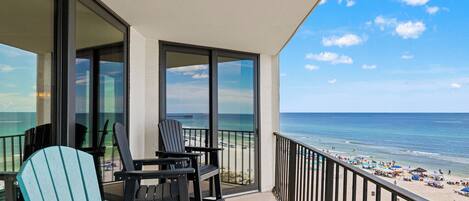 A balcony with two Adirondack chairs and a table, overlooking a beach with umbrellas and people.