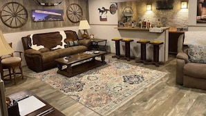 A living room with a brown leather sofa, a coffee table, and a patterned rug. There is a bar area with stools and a brick wall. - StockYards!  Walk 3 Blocks-Ranch House sleeps 8, TCU close by (Ft Worth)