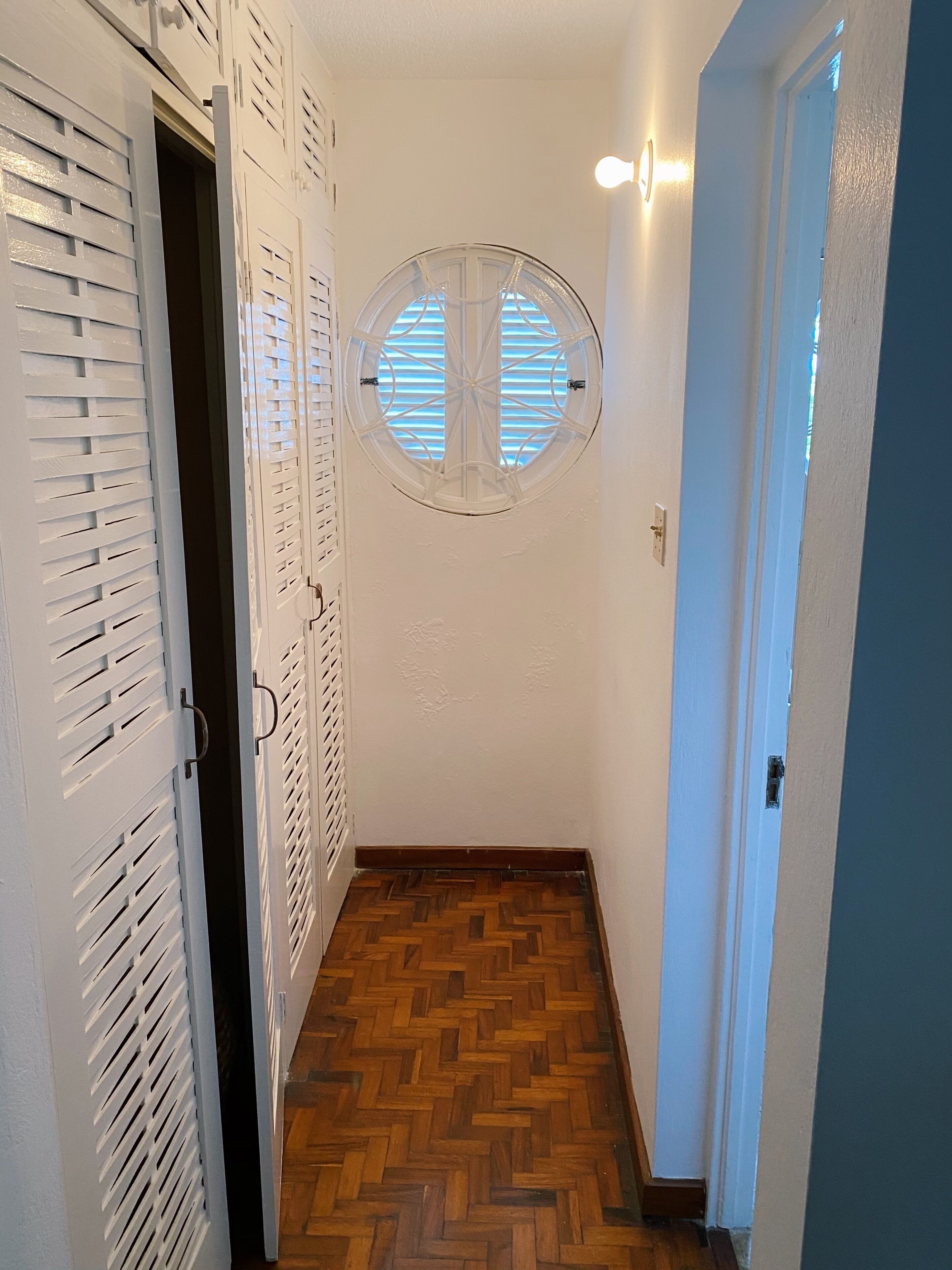 Een gang met een houten visgraatvloer, witte muren en een rond raam met jaloezieën.