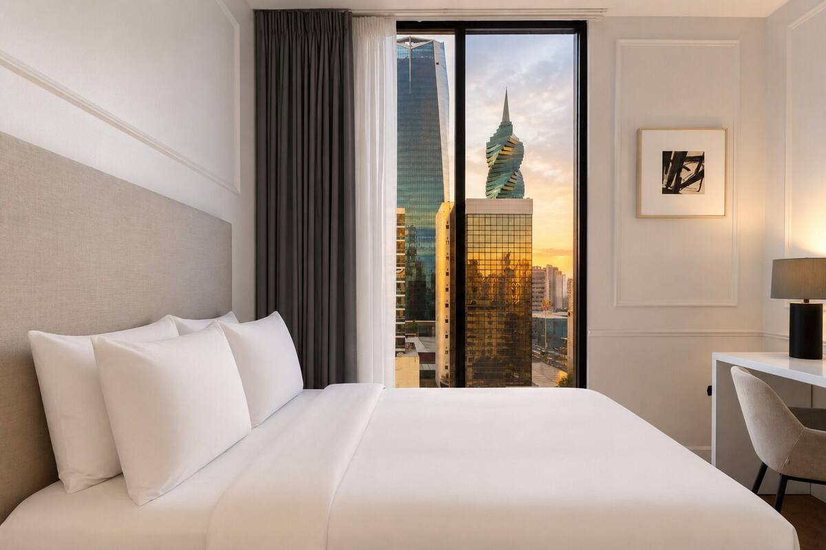 Une chambre d’hôtel avec un grand lit, un bureau, une chaise, une vue sur la ville et un tableau encadré au mur.