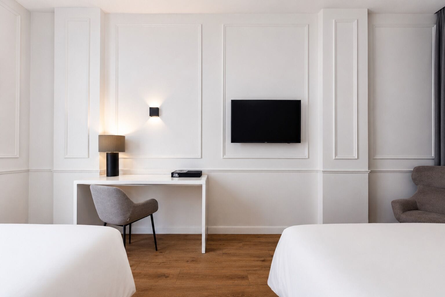 A hotel room with two beds, a desk with a chair, a TV mounted on the wall, and a lamp.