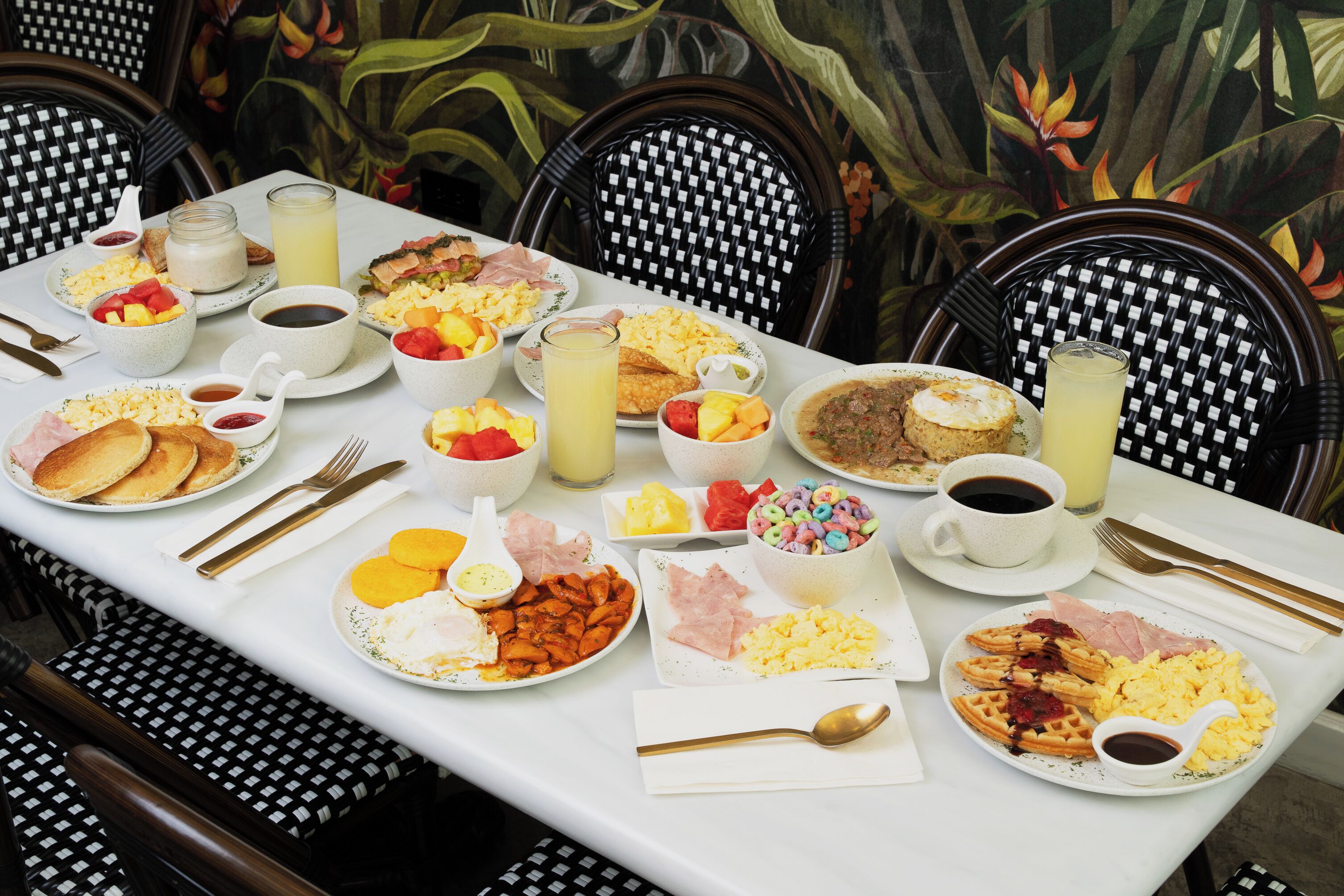 A table set with breakfast items, including pancakes, eggs, and waffles, with a tropical mural in the background.