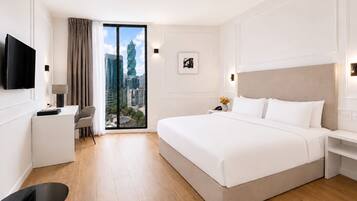 A modern hotel room with a large bed, a desk, a chair, a TV, and a view of the cityscape.