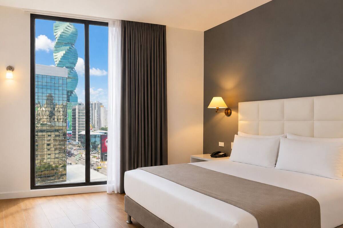 Une chambre d’hôtel avec un grand lit, une table de chevet, une lampe et une vue sur le paysage urbain, visible à travers une grande fenêtre.