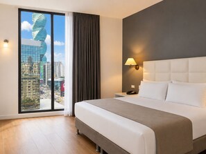 Hotelkamer met een groot bed, een nachtkastje, een lamp en uitzicht op een stadsgezicht door een groot raam.