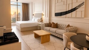 Habitación de hotel con cama, sofá, mesa de centro, escritorio y una ventana grande con vistas a la ciudad.