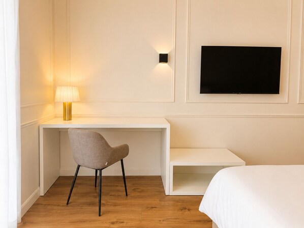 Une chambre d’hôtel avec un bureau blanc, une chaise grise, un lit, un téléviseur fixé au mur et une lampe sur le bureau.