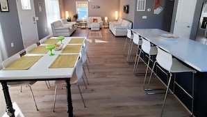 A spacious dining area with a long table and chairs, a kitchen area with a counter and stools, and a living room with sofas and a TV. - Private Retreat Bungalow-One Level, 2 blocks from Beach w/Porch & Deck (Colonial Beach)