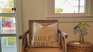 En træstol med pude, hvor der står "SURF SHACK", i et rum med et sivbord og en vase med en plante.