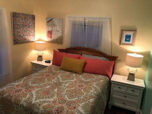 A bedroom with a bed, two bedside tables, a nightstand, a window with blinds, and a wall with a framed picture.
