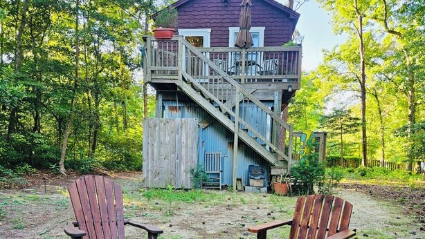 A rustic cabin with a deck, a fire pit, and Adirondack chairs in a wooded area.