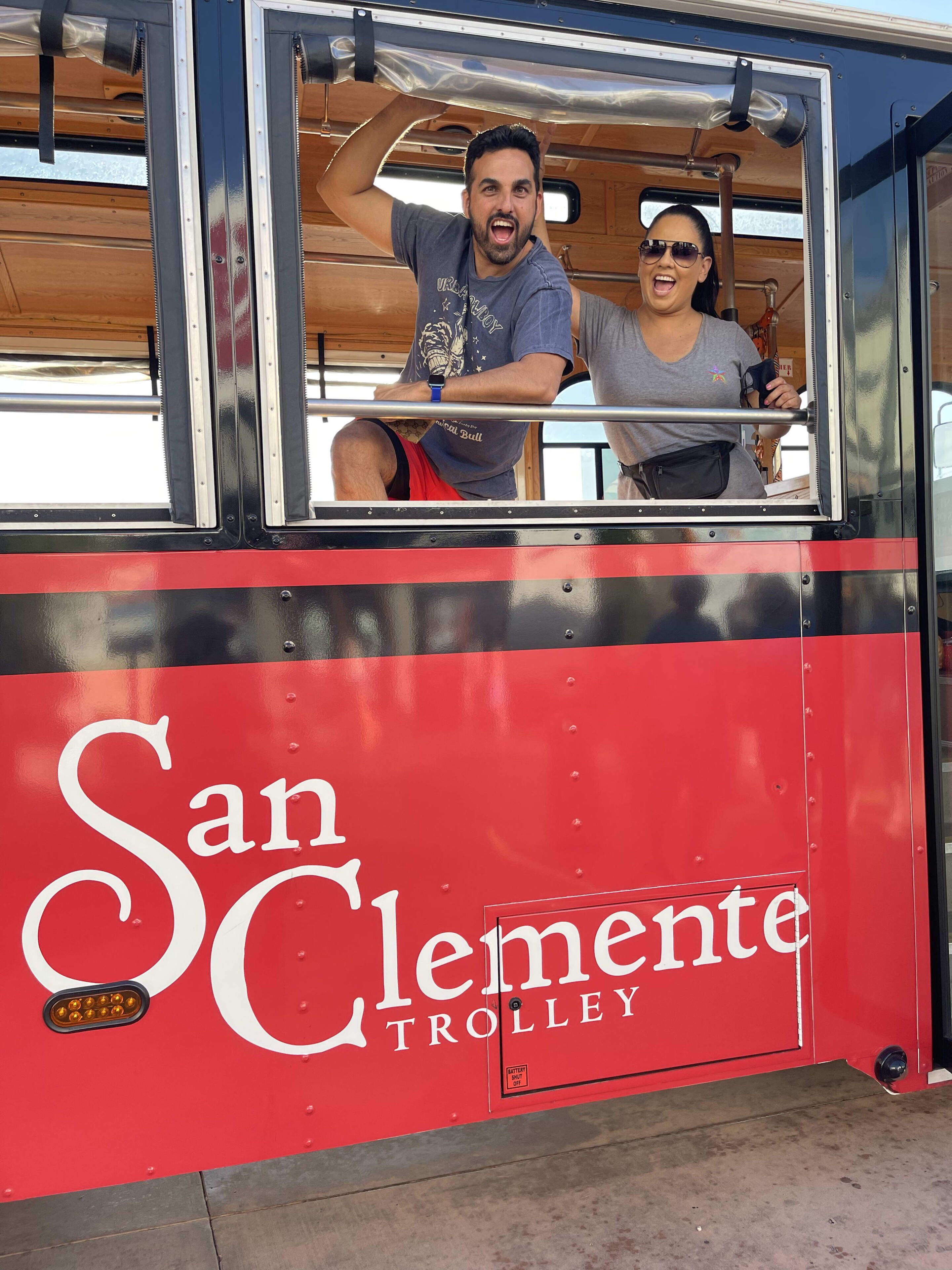 A red San Clemente Trolley with two people inside, windows, and a metal railing.