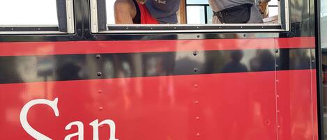A red San Clemente Trolley with two people inside, windows, and a metal railing.