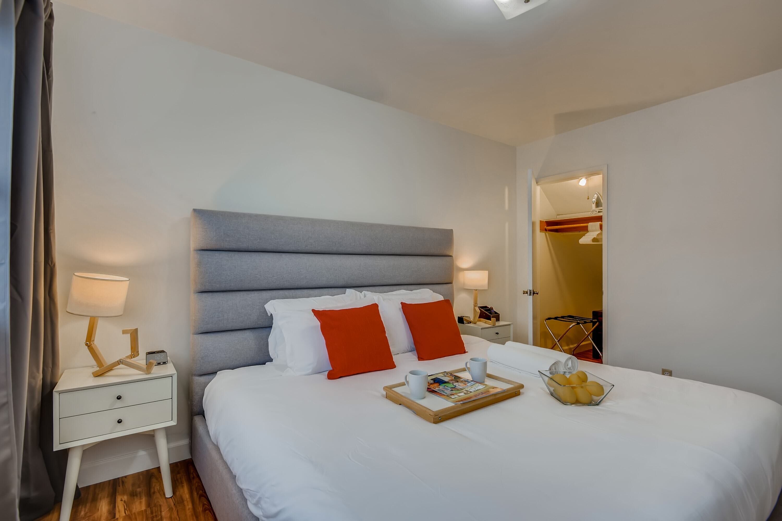 A neatly made bed with white and orange pillows, a bedside table with a lamp, and a tray with a book and fruit.