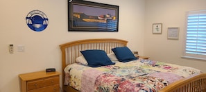A bedroom with a wooden bed, a colorful quilt, two blue pillows, a framed painting, a wall clock, and a window with blinds.