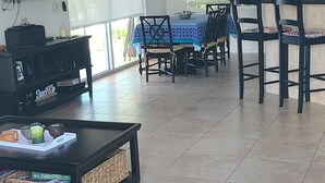 Amplia sala de estar con televisor, ventiladores de techo, comedor con sillas y una mesa, y cocina con armarios.