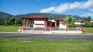 A building with a red roof and white walls, surrounded by a white fence, with a road in the foreground and mountains in the background. - Villa Dorada Jarabacoa (Jarabacoa)