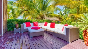 Una terraza de madera con un sofá seccional blanco y cojines rojos, rodeada de exuberante follaje y palmeras.